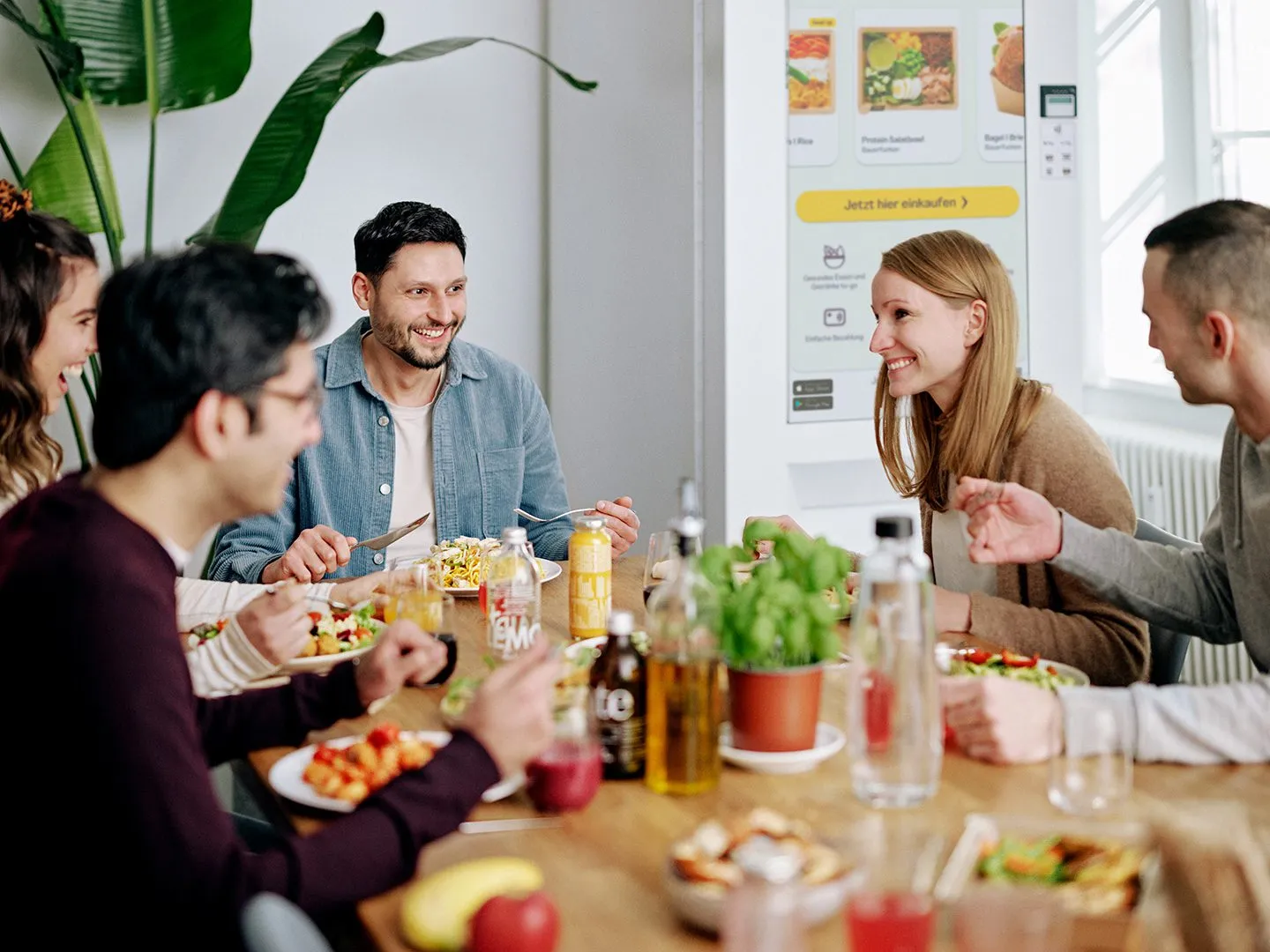 Eine Büroküche, in der Mitarbeiter zu Mittag essen, glücklich und engagiert aussehen, mit einem intelligenten Verkaufsautomaten im Hintergrund sichtbar.